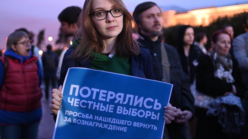 A woman with brown hair and wearing glasses holds up a protest sign.