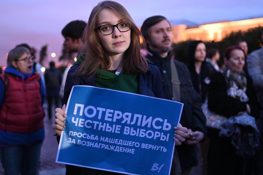 A woman with brown hair and wearing glasses holds up a protest sign.