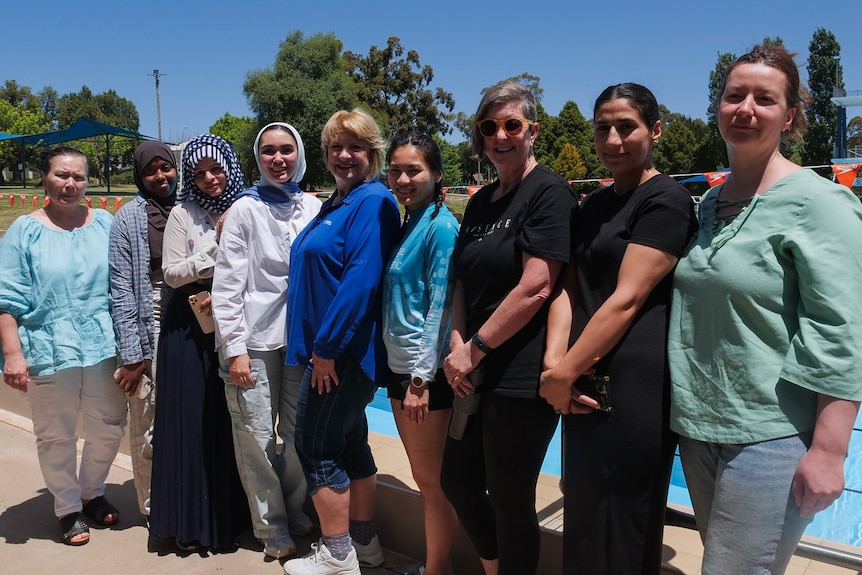 Nine women stand together in front of an outdoor 50 metre public pool.