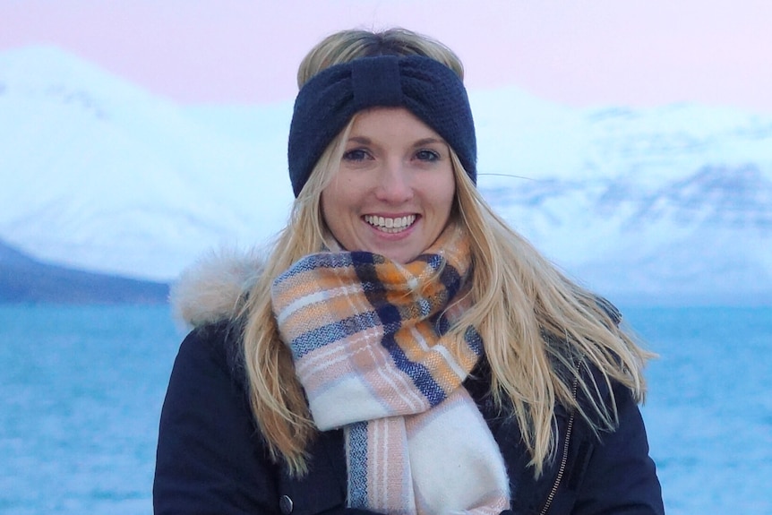 A blonde woman wearing a scarf and head band smiles in front of giant snowy mountain in the background