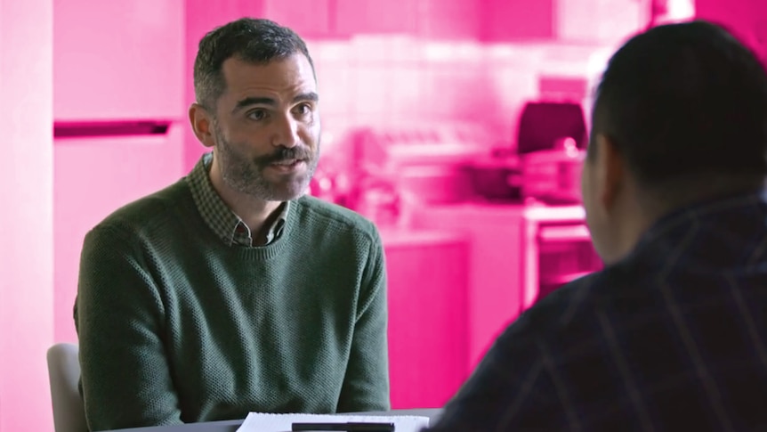 A man with a short beard talks to another man across a table, seen over his shoulder, a notepad and pen between them