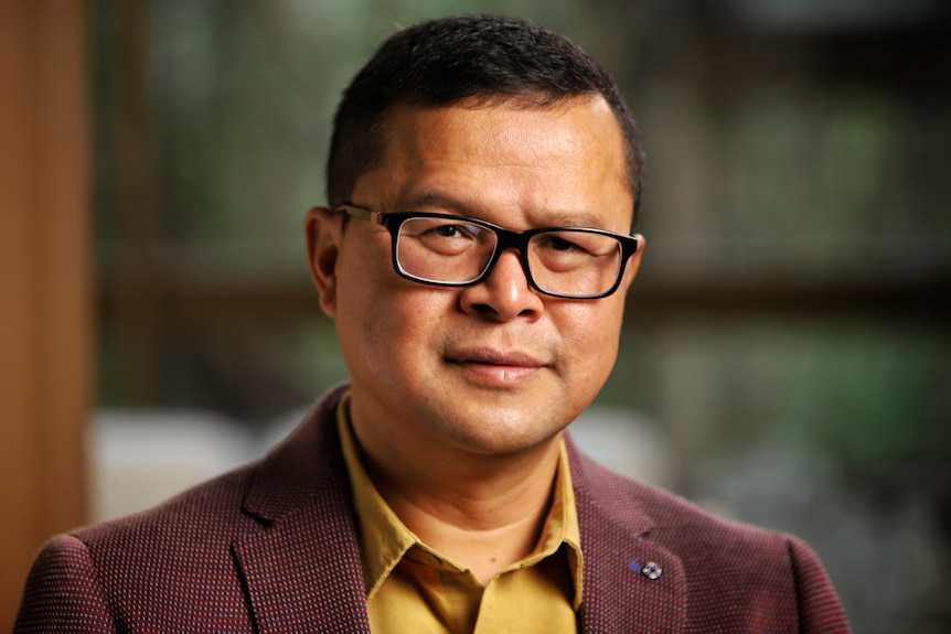 Dr Dicky Budiman is a Queensland-based epidemiologist. He is wearing glasses, a brown jacket and mustard shirt