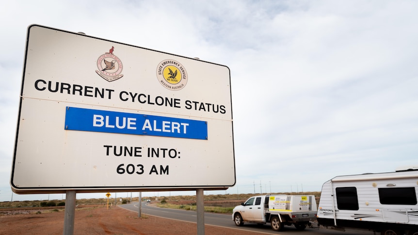 A blue alert sign on the outskirts of Port Hedland, with a departing caravan in the background.