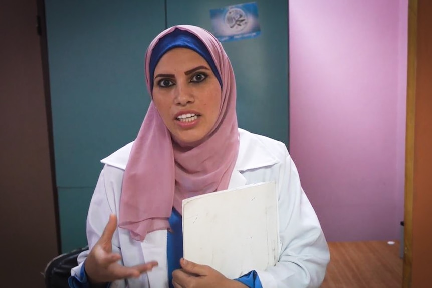 A woman wearing a headscarf and a doctors coat talks while holding a manilla folder.
