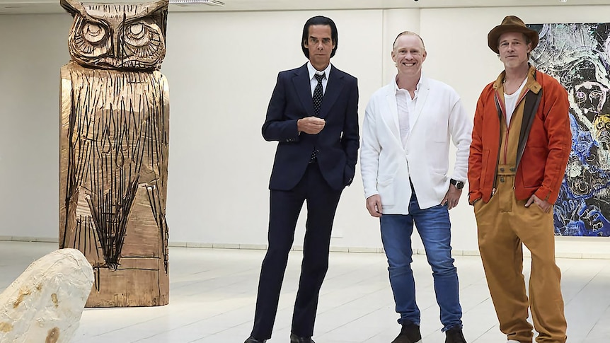 Nick Cave and Brad Pitt pictured in an art gallery next to a large human-sized sculpture of an owl.