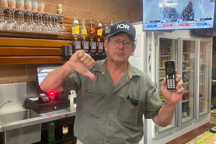 A male publican holds up a phone in one hand, with a thumbs down gesture in the other