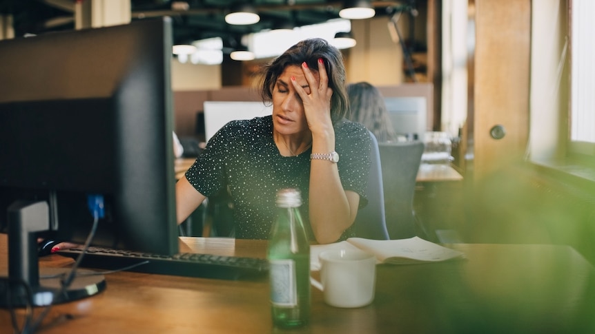 A multiracial woman sitting at a desk, looking tired, with one hand raised to her forehead