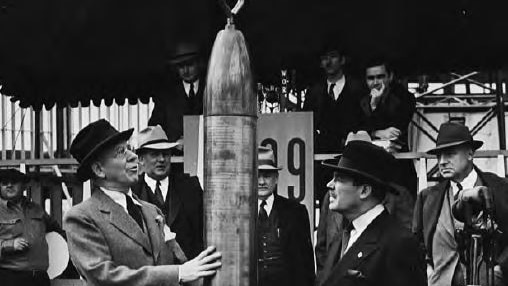 A black and white photo of two men in hats standing either side of a time capsule that looks like a rocket