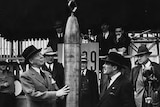 A black and white photo of two men in hats standing either side of a time capsule that looks like a rocket