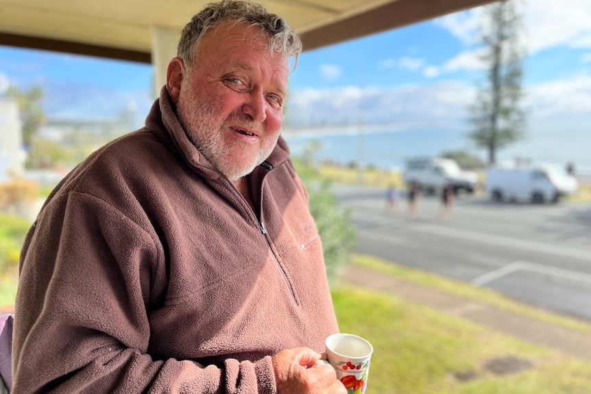 older bearded man holding coffee standing on verandah with ocean behind