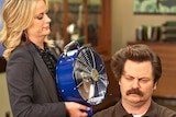 Amy Poehler, playing Leslie in Parks and Recreation, holds a blue fan up to the face of Ron, played by Nick Offerman