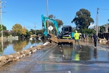 A crane works to repair flooding damage on a road. A wall of sandbags blocks flooding.