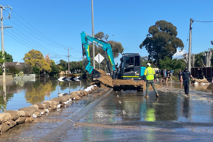 A crane works to repair flooding damage on a road. A wall of sandbags blocks flooding.