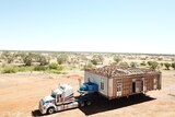 Truck tows trailer with house on a dusty outback road.