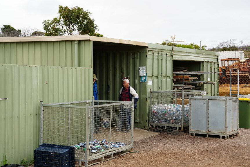 Un hombre está parado en la puerta abierta de un cobertizo, con cajas de contenedores a cada lado. 