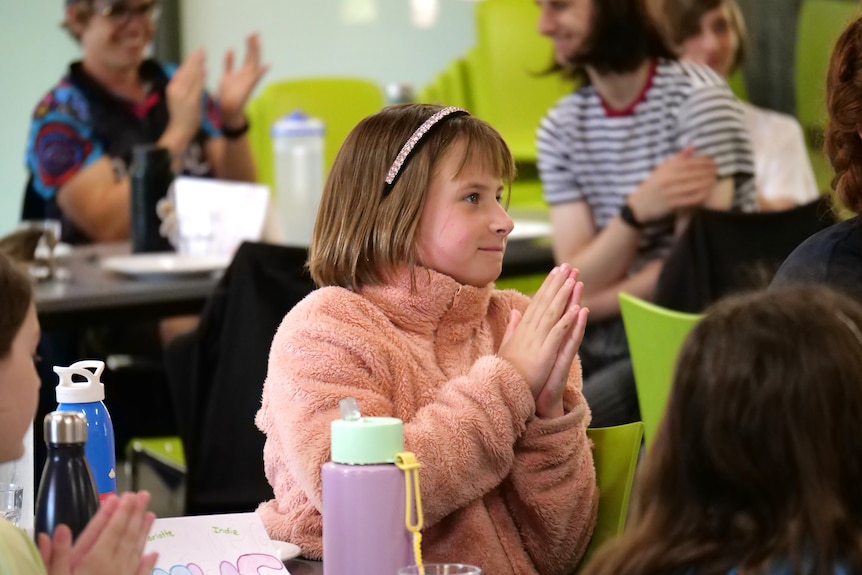 A girl in a pink jumper sits in a dining room and claps, looking across from the camera to the front of the room.