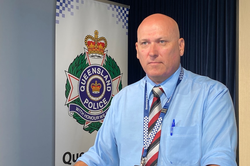 a man wearing a blue shirt and a striped tie in front of a Queensland Police sign