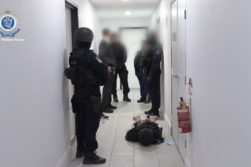 Multiple police officers in protective clothing stand in a narrow hallway while a handcuffed man lies face down on the floo