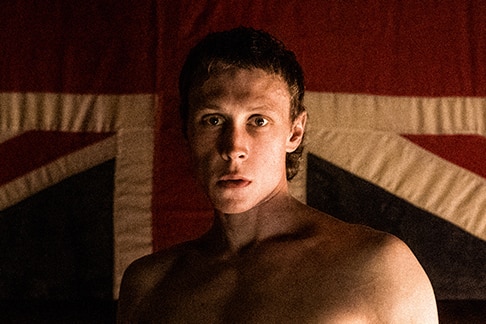 A topless man with short cropped hair stands in front of large Union Jack flag hanging from ornate patterned wallpaper.