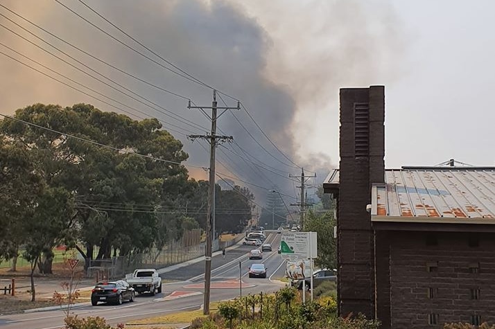 Smoke billows at the end of a road into the sky surrounded by houses and cars.