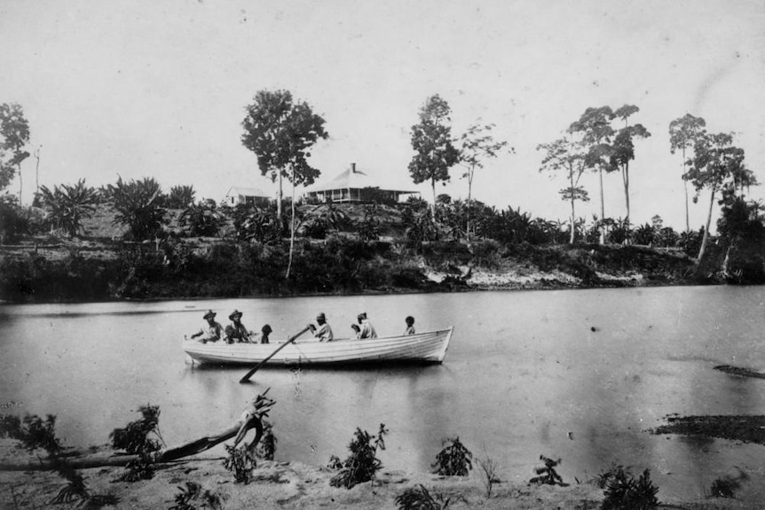 A black and white image showing a wooden boat full of people rowing down a river, a building is visible in the background.