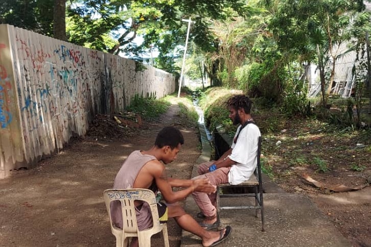 Two men sitting on chairs on a dirt road.