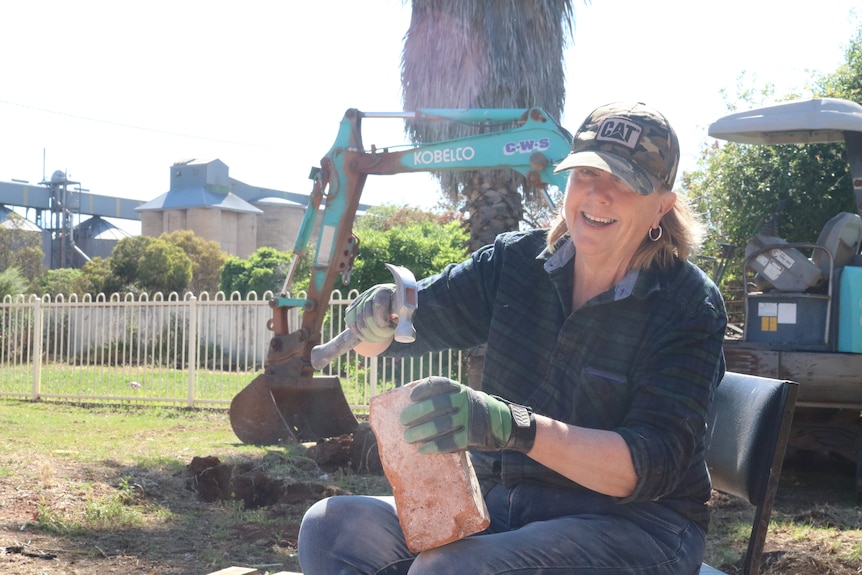 A woman sitting down and chipping cement of bricks with a backhoe in the background.