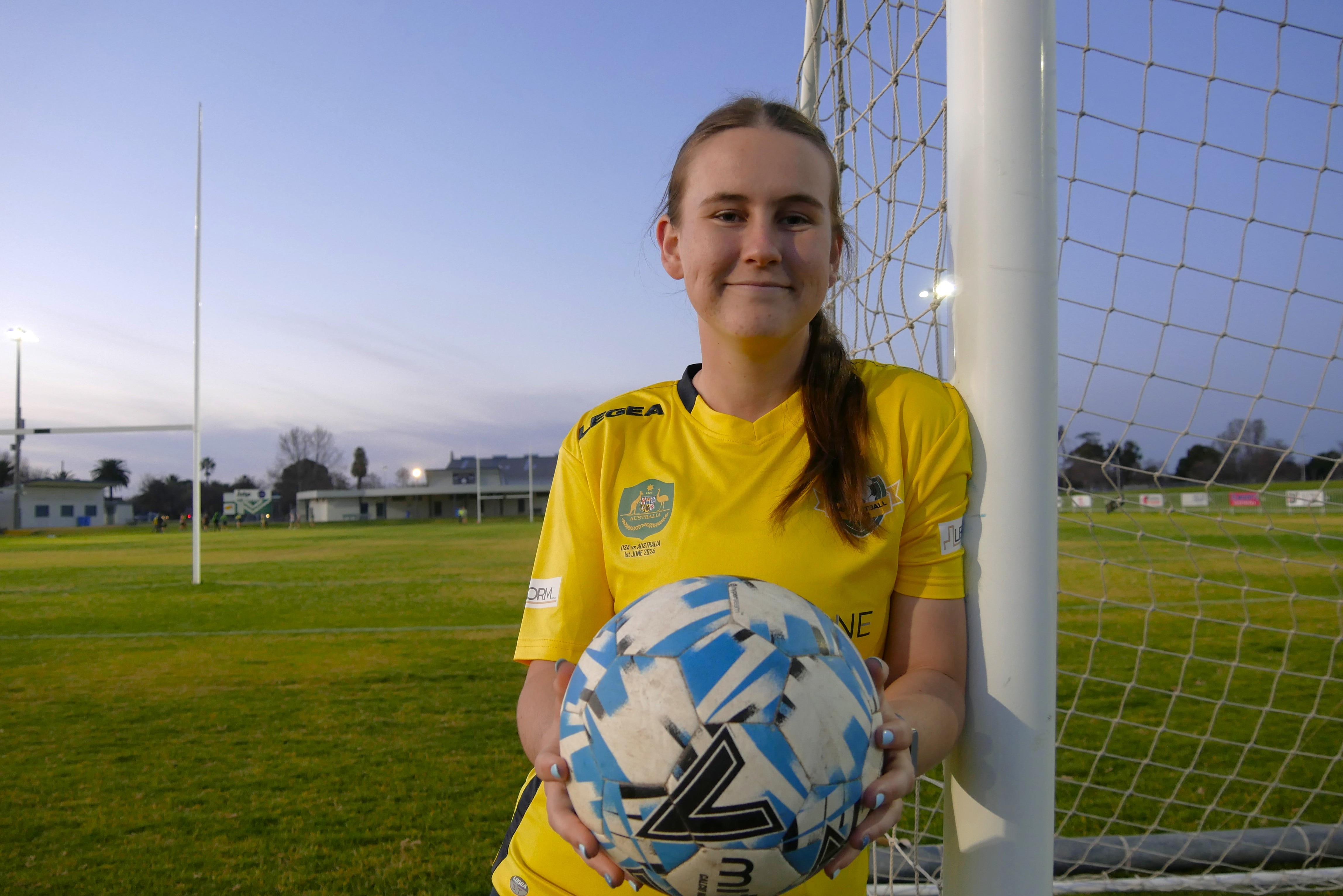 A teenage girl holds a football and wears a green and gold jersey, leaning against a goal.