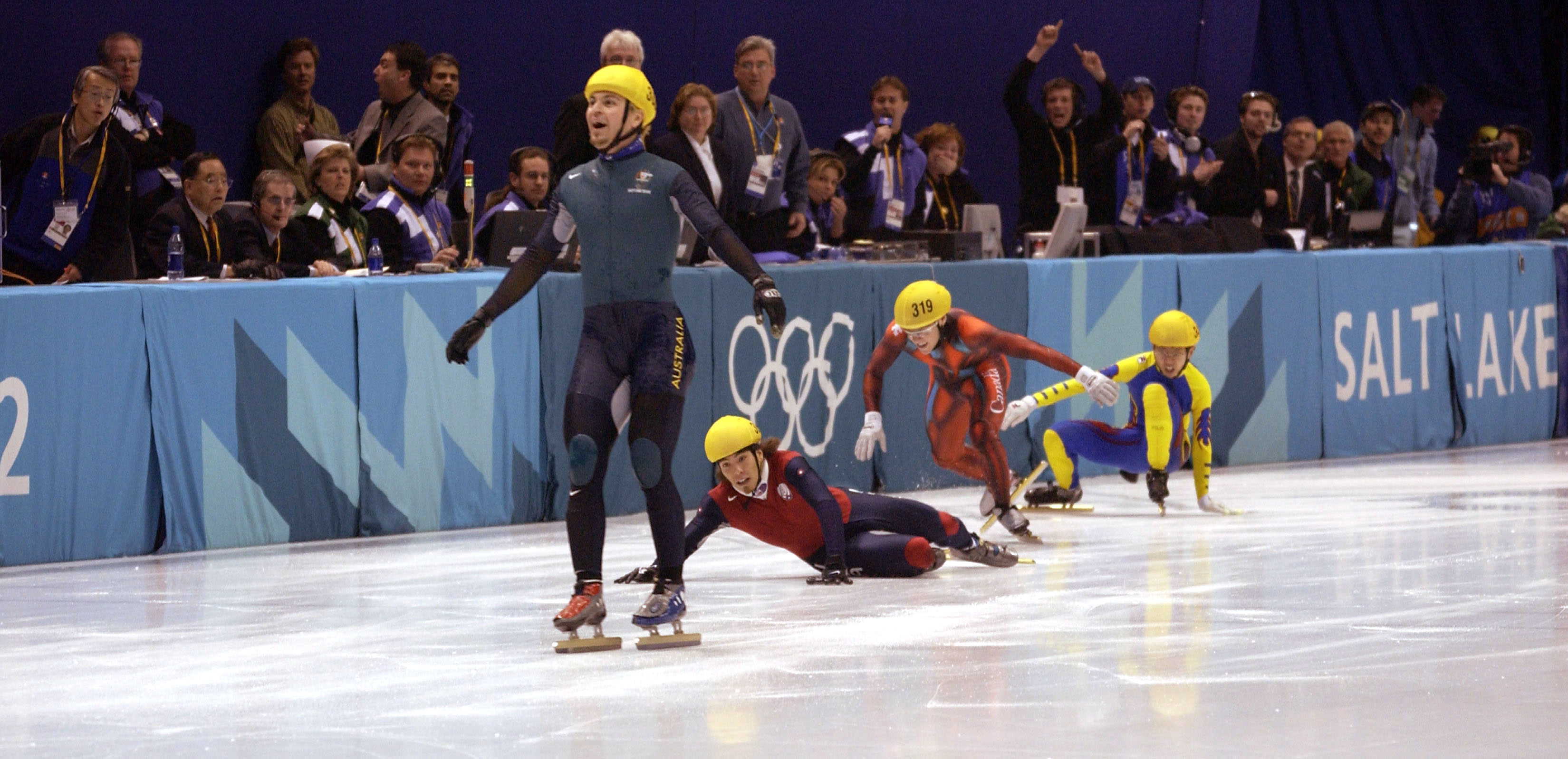Steven Bradbury desliza sobre a linha com patinadores no chão atrás dele