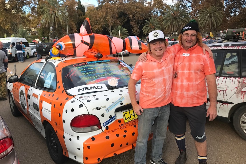 Two men in colourful costumes with rally cars behind them.