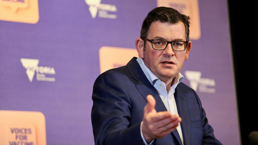 Daniel Andrews gestures with a hand, with a serious expression on his face, at a podium in front of a purple backdrop.