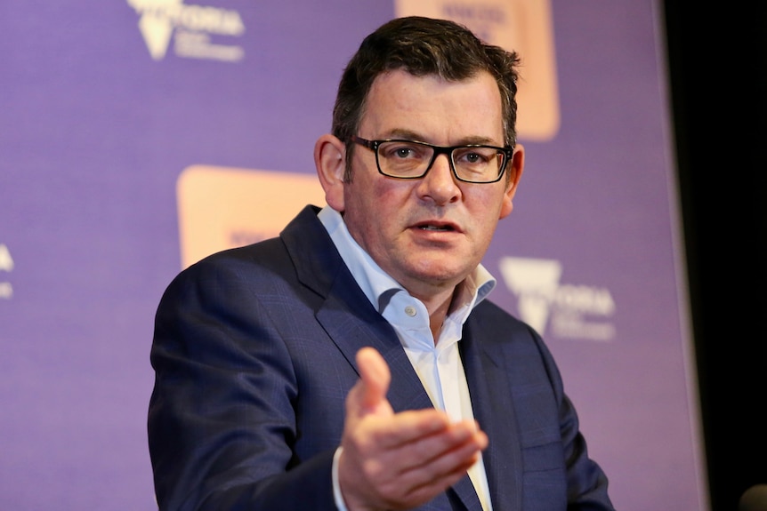 Daniel Andrews gestures with a hand, with a serious expression on his face, at a podium in front of a purple backdrop.