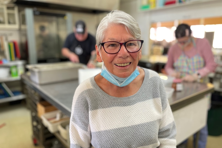 An older woman with grey hair, smiling inside a community kitchen.