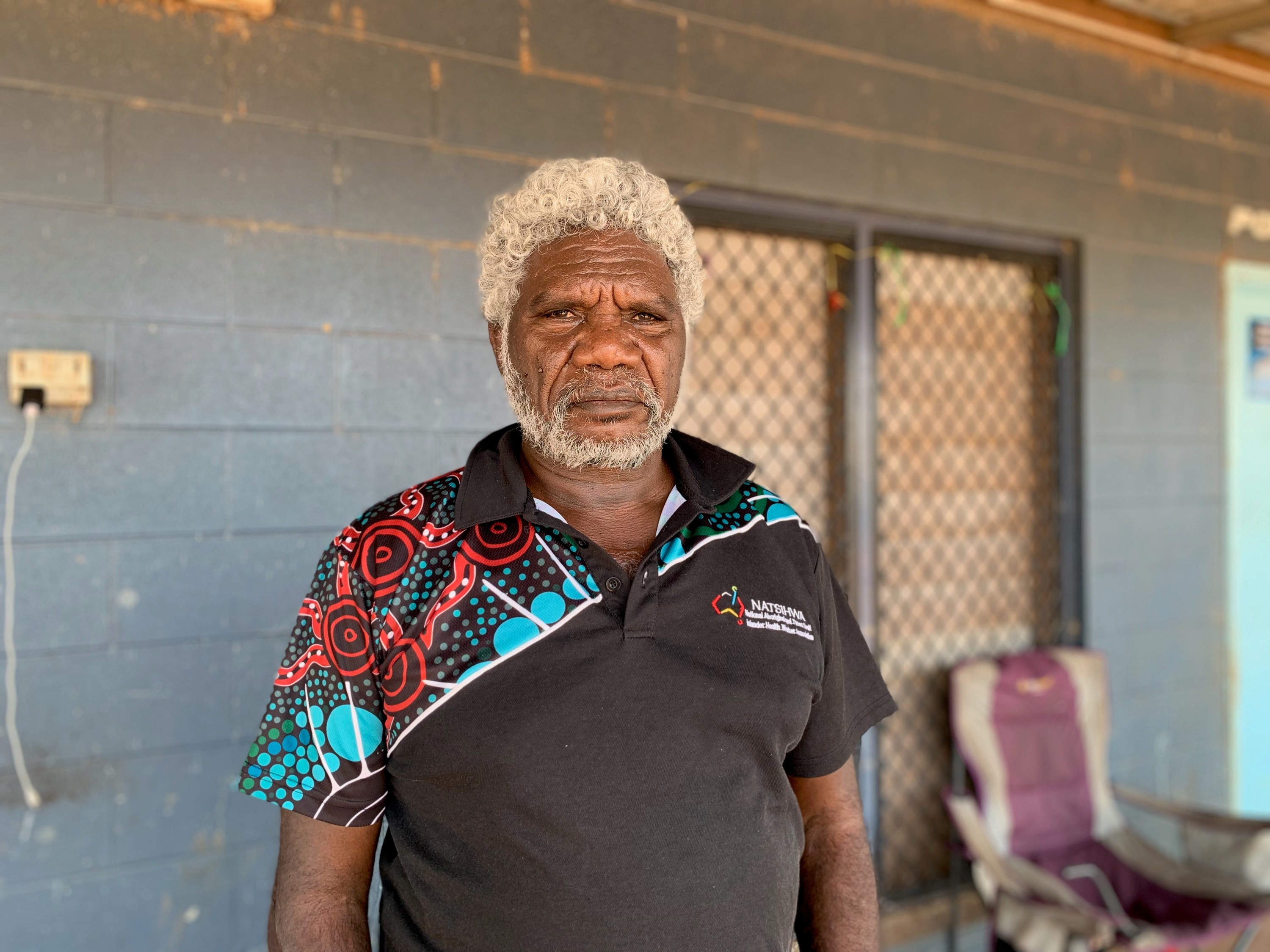 Aboriginal man, gray curly short hair and beard standing in front of house, black polo shirt on, sombre expression.