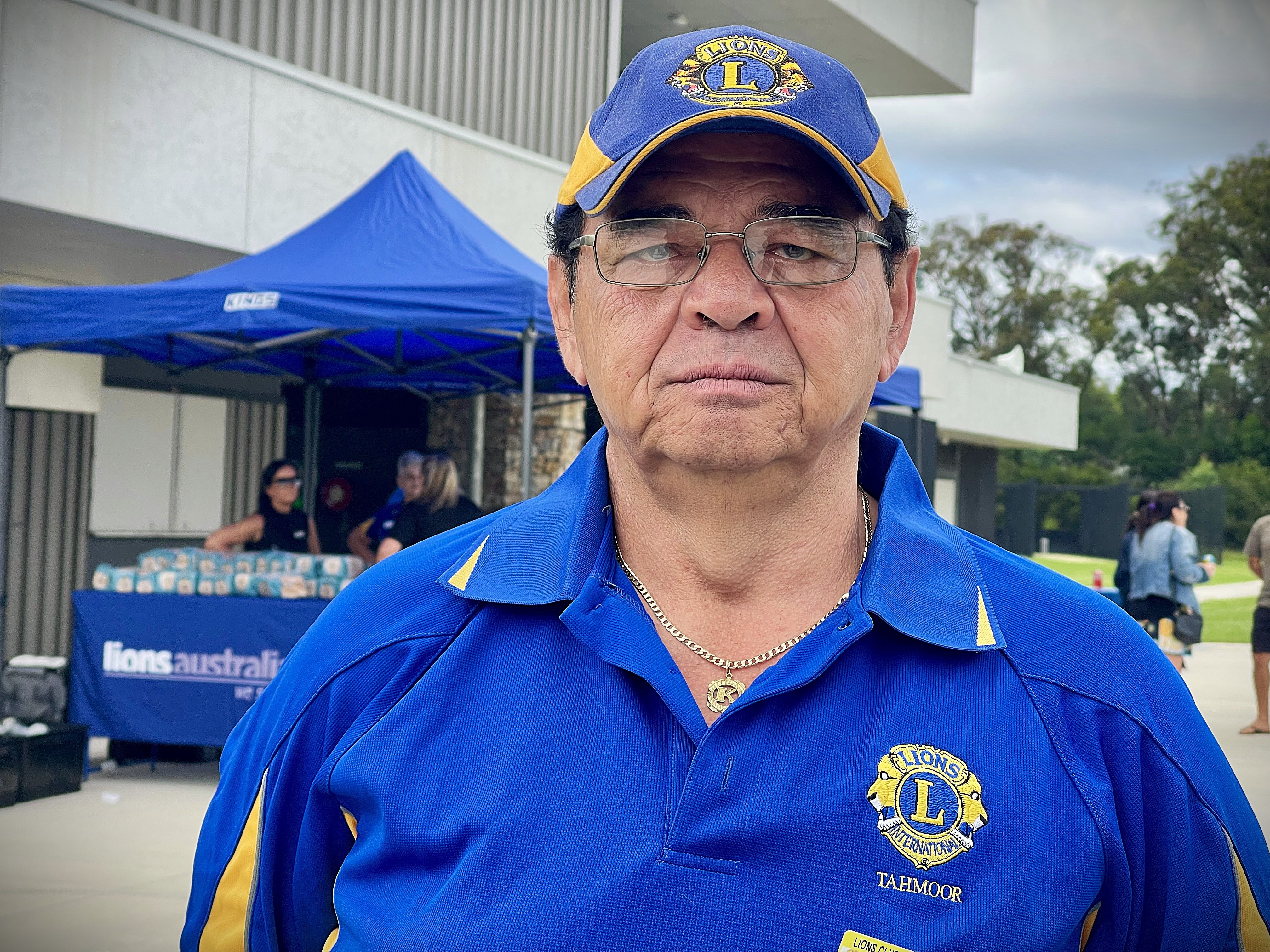 Hombre con camisa de león azul y gorra frente al puesto de barbacoa.