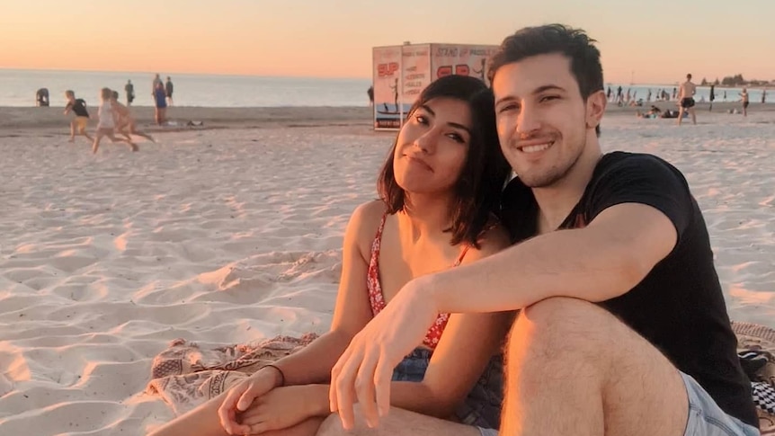 A man and woman sit next to each other on the sand at sunset, smiling.