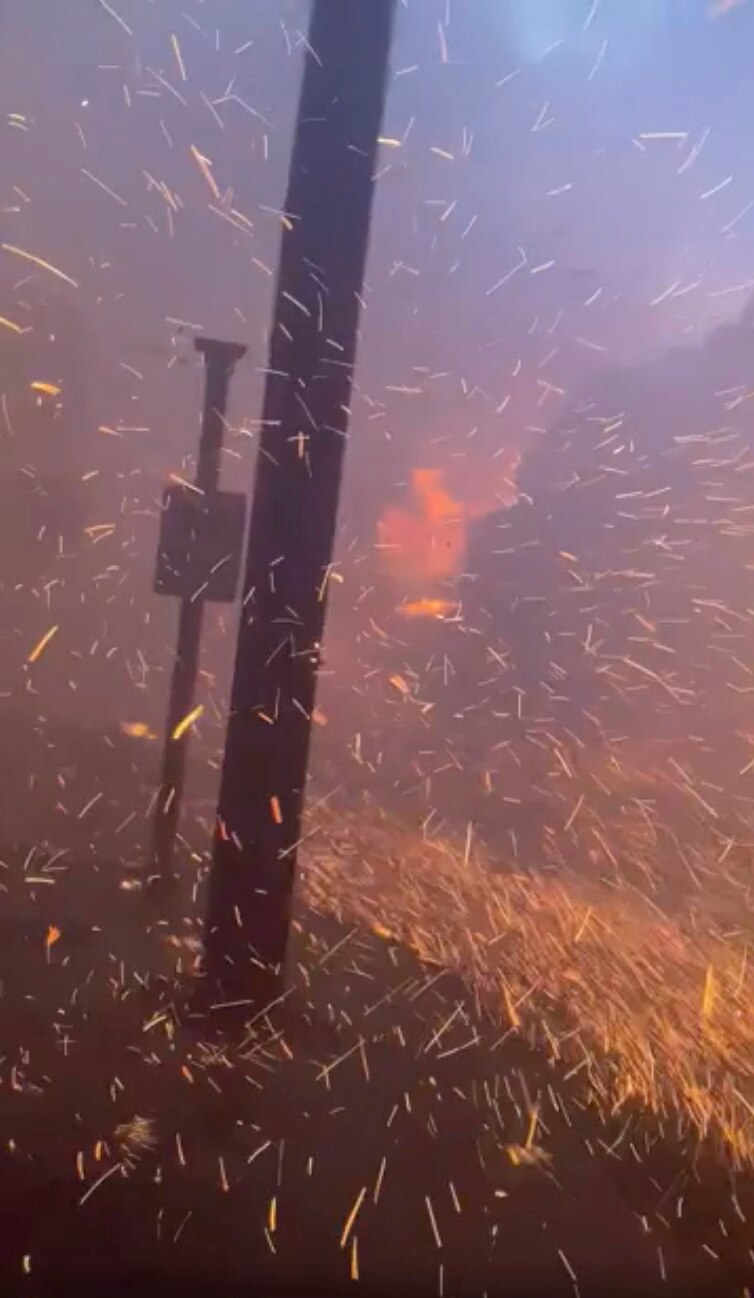 Glowing red and orange embers fill the frame, a power pole and street sign barely visible in smoky, fiery conditions