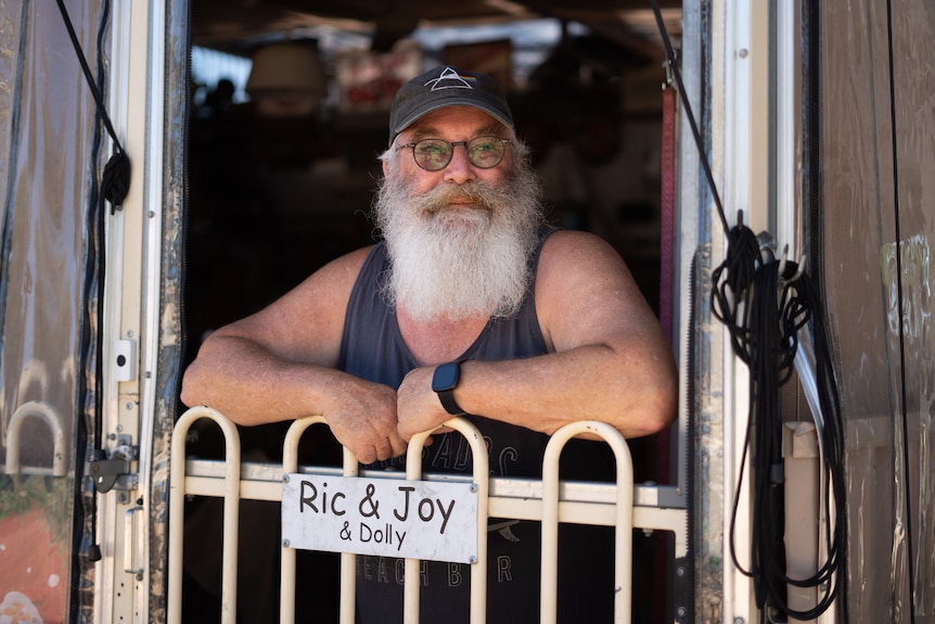 A man with a bushy grey beard smiles, he wears a dark tank top and a dark hat. He smiles gently leaning over a gate