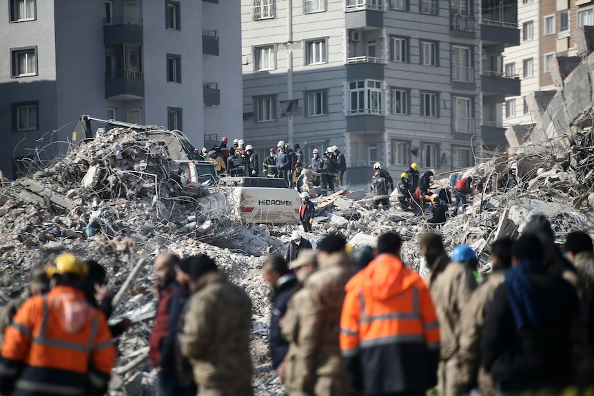 A view from a distance of a pile of rubble from a collapsed building, with earth movers and workers looking for survivors.