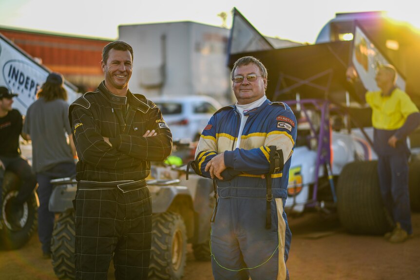 Two men with their arms crossed standing in front of sprint cars in uniform