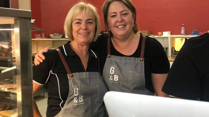 Two women in matching overalls stand smiling with their arms around each other in a cafe.