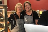 Two women in matching overalls stand smiling with their arms around each other in a cafe.