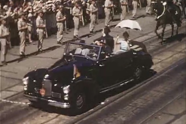 The Queen driving along in a convoy through Brisbane city flanked by horses and soldiers.