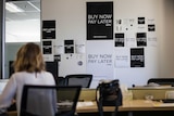 A woman sits at a desk in an office with "buy now, pay later" posters behind her