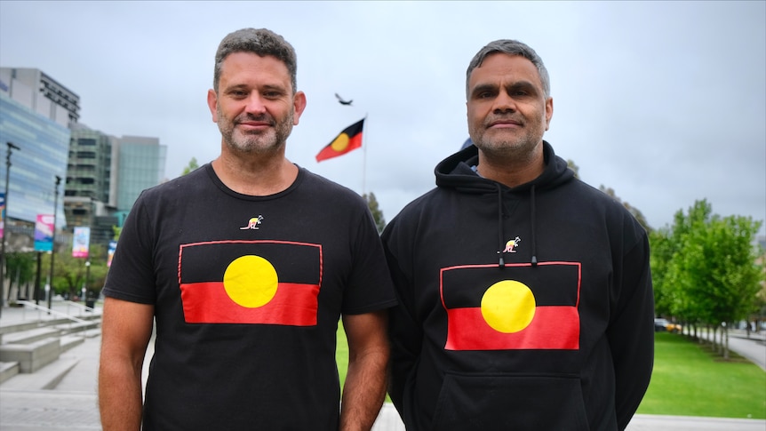 Two smiling men wearing black tops with the Aboriginal flag on it. An Aboriginal flag can also be seen in the background