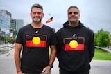 Two smiling men wearing black tops with the Aboriginal flag on it. An Aboriginal flag can also be seen in the background