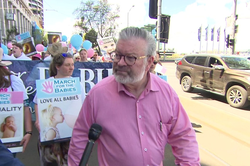 a man in a pink shirt with grey hair and a crowd holding signs behind him.