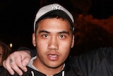 A young man in a black hoodie and grey baseball cap making a hand gesture standing next to a friend pointing at him.