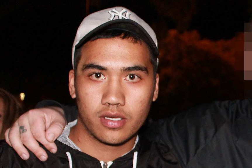 A young man in a black hoodie and grey baseball cap making a hand gesture standing next to a friend pointing at him.