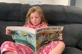 A girl in pink pyjamas sits crossed-legged on a lounge reading a book.